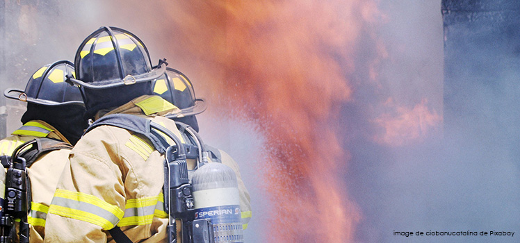 Trois pompiers interviennent pour maîtriser un incendie en projetant de l'eau sur de grandes flammes, au milieu d'une épaisse fumée.